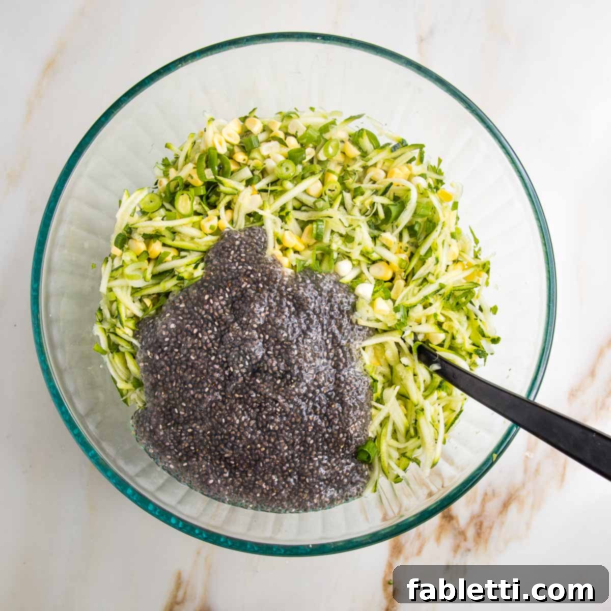 Crispy Zucchini Corn Patties 9 A large glass bowl containing a mixture of shredded zucchini and corn, with two golden-brown chia eggs gently poured over the top, ready to be mixed into the vegetable fritter batter.