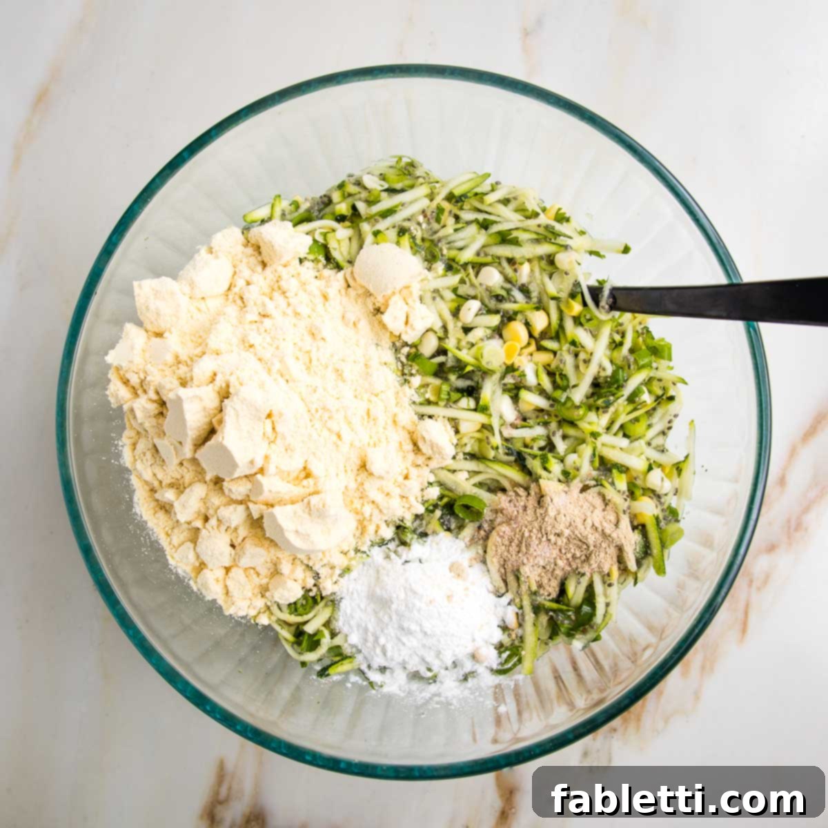 Crispy Zucchini Corn Patties 10 A large glass bowl holding the wet mixture for zucchini corn fritters, with a layer of dry ingredients—cornmeal, baking powder, salt, and pepper—piled on top, ready to be incorporated into the batter.