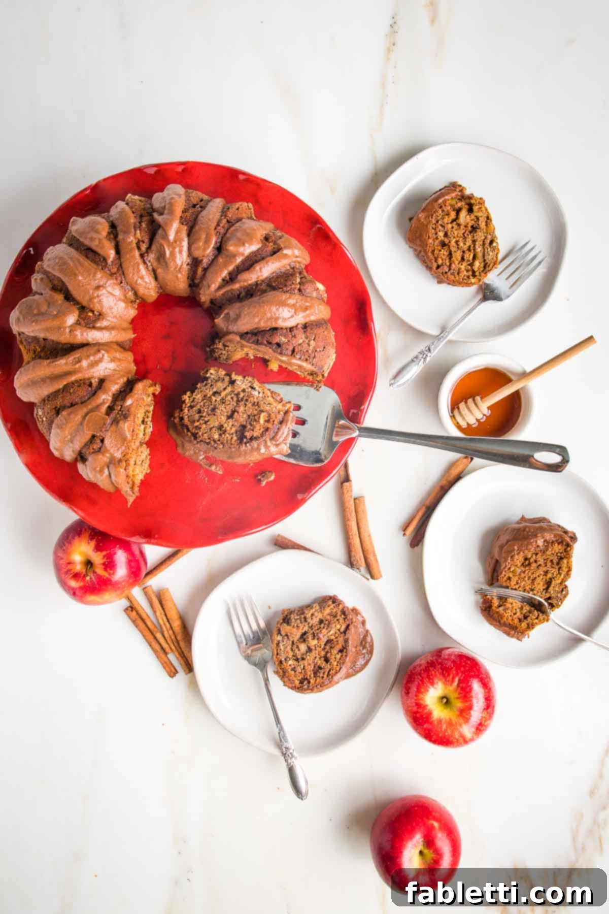 Spiced Apple Bundt Delight 2 Red pedestal with apple cake drizzled with chocolate sauce. Small plates with pieces of apple cake, a dish of honey and cinnamon sticks on the counter.