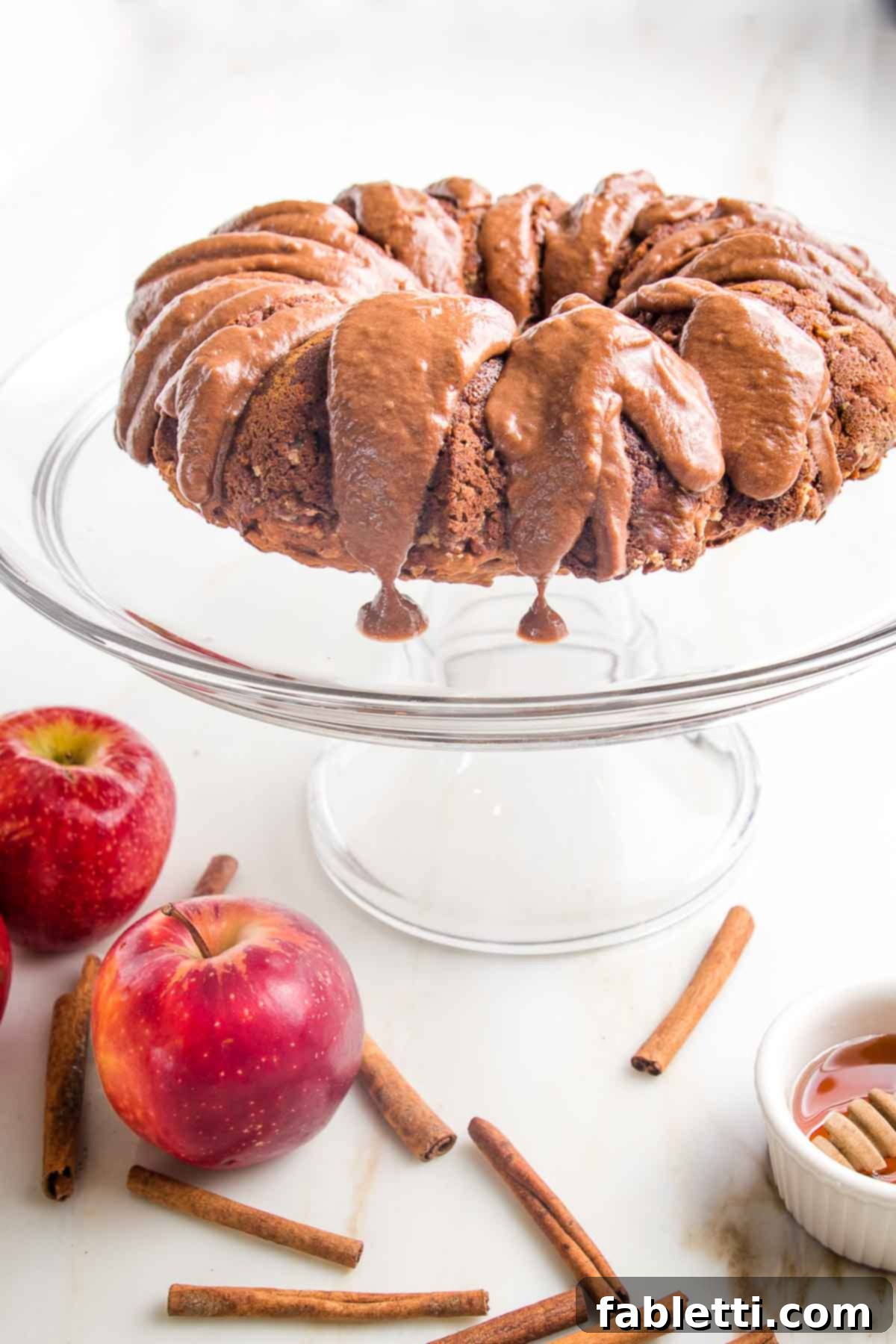 Spiced Apple Bundt Delight 13 Apple bundt cake with chocolate drizzle dripping down the side. The cake is on a glass pedestal and there are red apples and cinnamon sticks below.