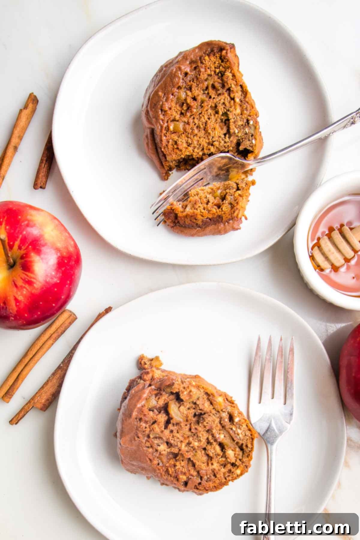 Spiced Apple Bundt Delight 14 Slice of apple honey cake on a plate with a fork cutting through. Another slice on a plate shows the chocolate frosting. Whole apples, cinnamon sticks and a bowl of honey are in the background.