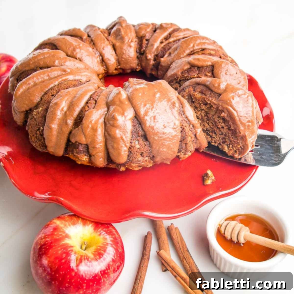 Spiced Apple Bundt Delight 15 Apple bundt cake with chocolate drizzle on a red cake stand, surrounded by whole apple, cinnamon sticks and honey.