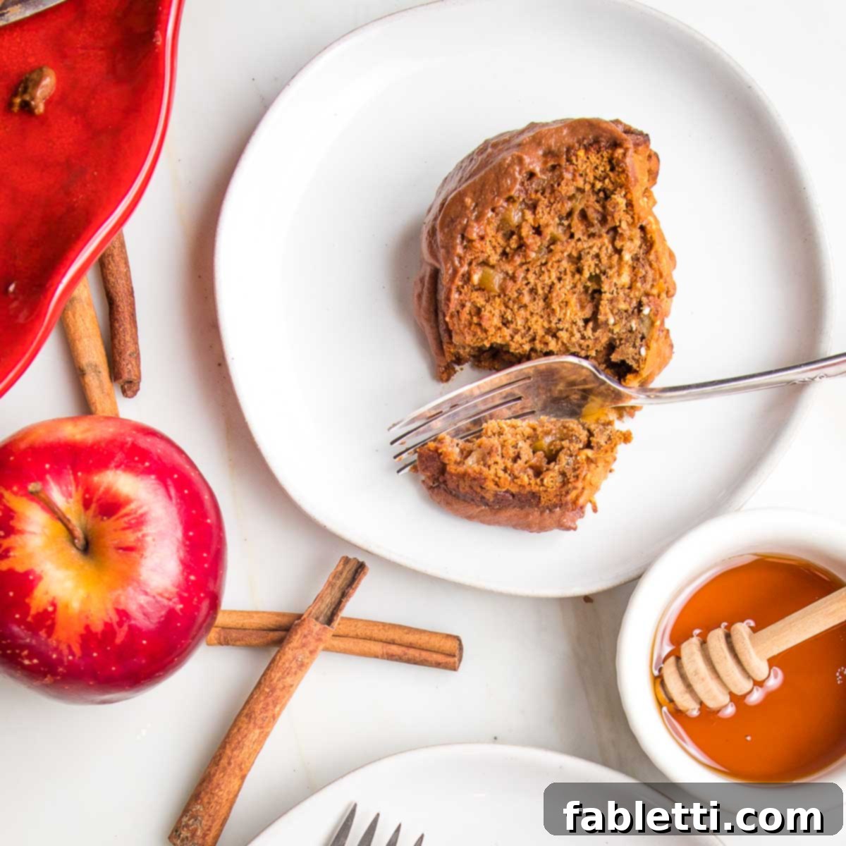 Spiced Apple Bundt Delight 4 A slice of apple honey cake with chocolate glaze on a white plate. Also shown: a whole red apple, cinnamon sticks and a dish of honey.