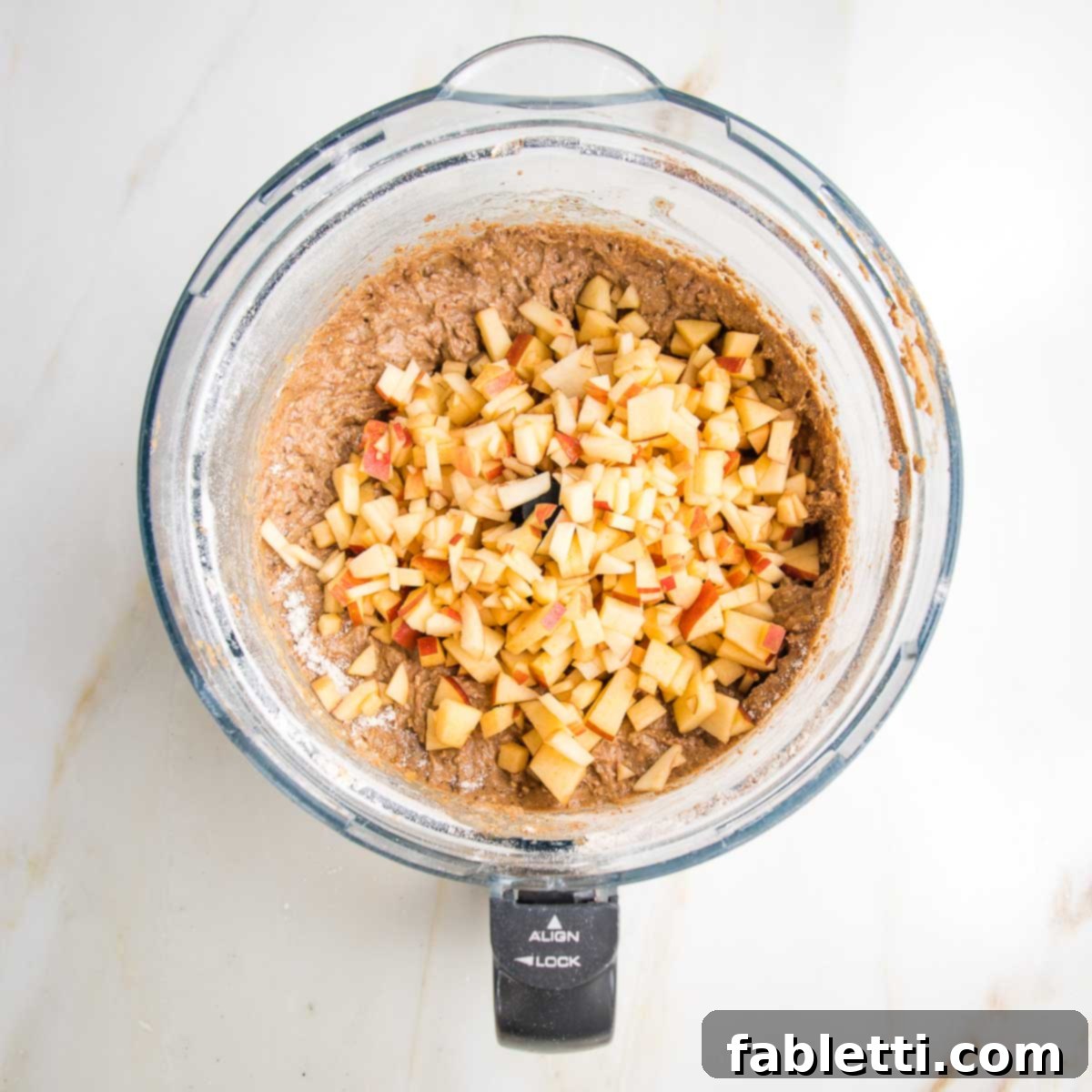 Spiced Apple Bundt Delight 7 Cake batter in the bowl of a food processor with chopped apple on top.
