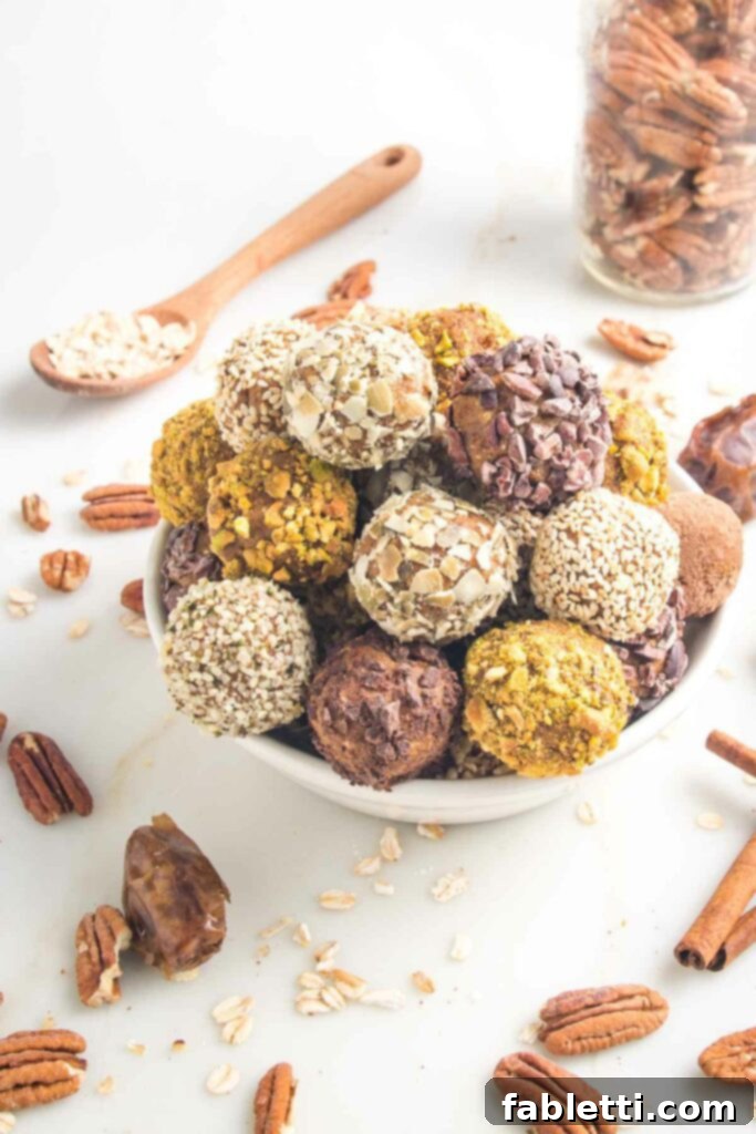 Pumpkin Spice Truffles 2 A bowl filled with Pumpkin Spice Bliss Balls, garnished with various crushed nuts and seeds. In the background, a spoon of oats and a jar of pecans are visible, with scattered whole nuts, dates, and cinnamon sticks in the foreground.