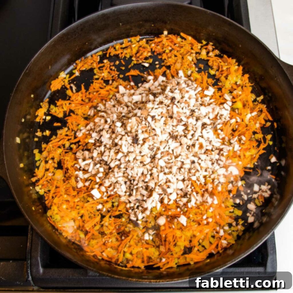 Hearty Vegan Bolognese over Squash Strands 10 A cast iron skillet with sautéed shredded carrots and other vegetables, topped with small diced mushrooms waiting to be mixed in and cooked.