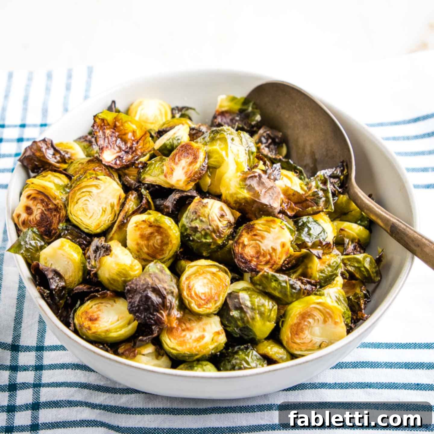 Honey Sriracha Glazed Crispy Roasted Brussels Sprouts 3 A rustic white bowl overflowing with golden, crispy brussels sprouts, set against a striped teal and white tea towel, highlighting their appealing texture.