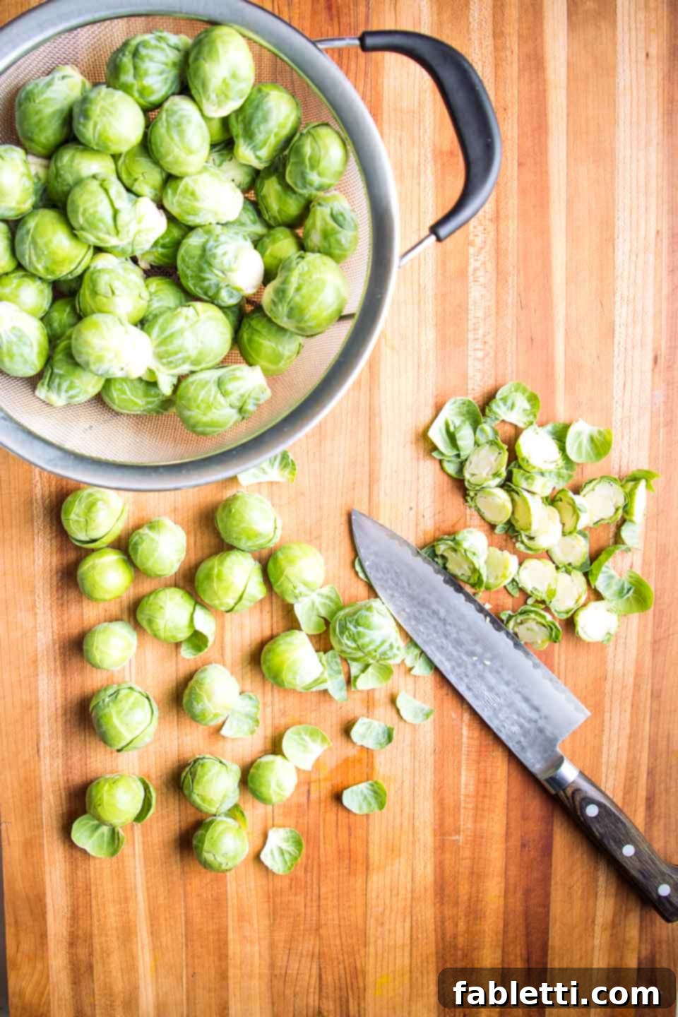Honey Sriracha Glazed Crispy Roasted Brussels Sprouts 4 A colander full of freshly washed Brussels sprouts next to a cutting board, where a sharp knife is trimming the ends of the sprouts, preparing them for halving.