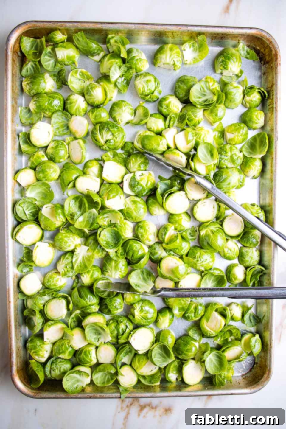 Honey Sriracha Glazed Crispy Roasted Brussels Sprouts 5 A large rimmed baking tray with halved Brussels sprouts being tossed with tongs, ensuring they are evenly coated with olive oil and seasoning before roasting.