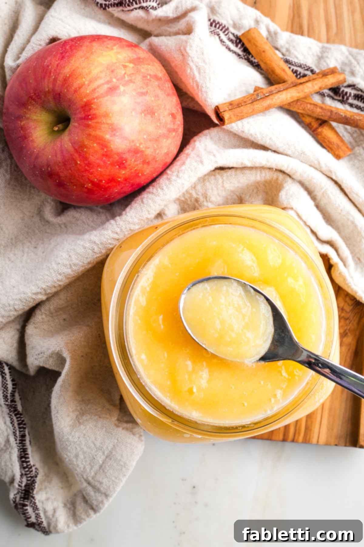 Jar of applesauce with an apple and cinnamon sticks in the background.