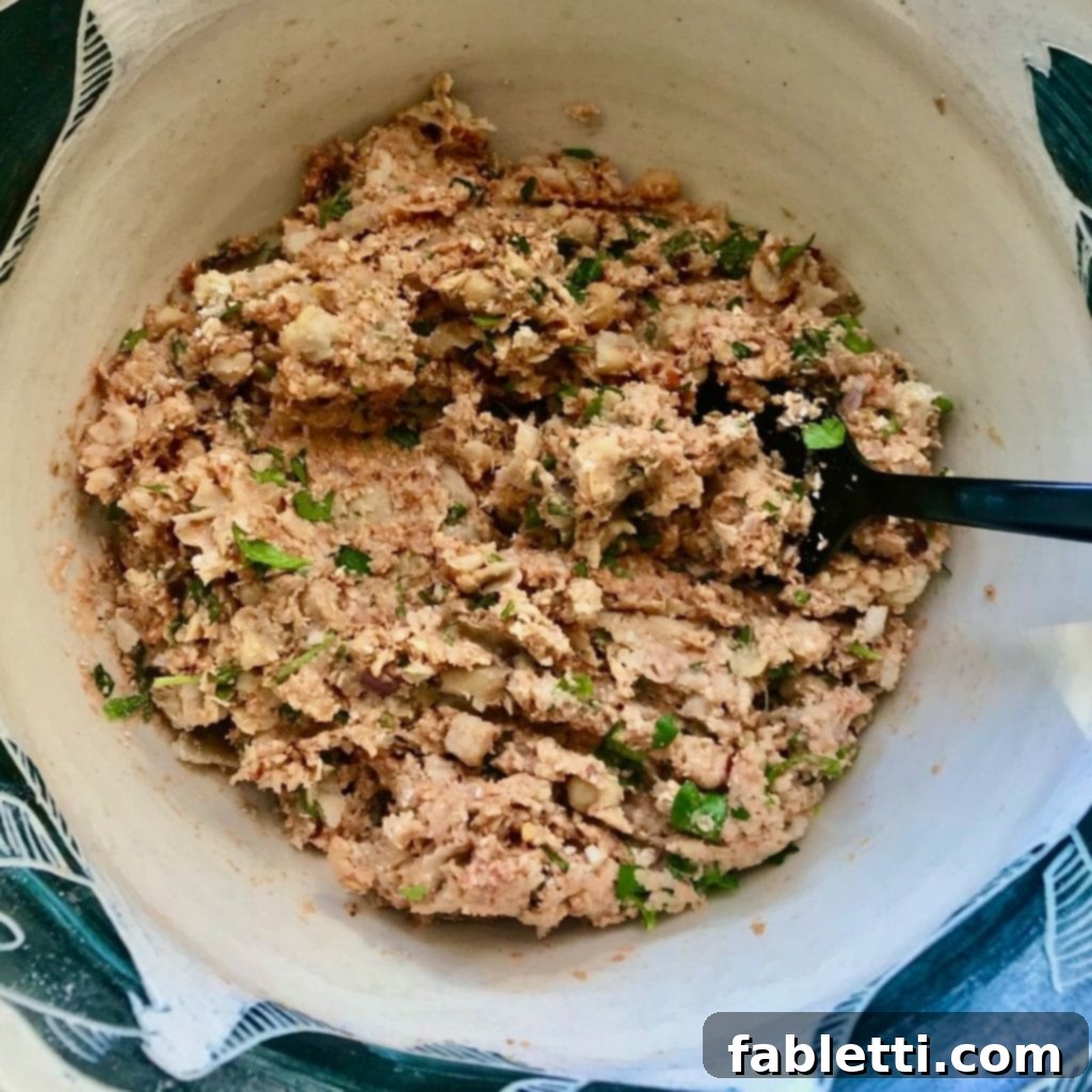 Beige bowl with a large black fork actively mashing together chickpeas, rich tomato paste, vibrant fresh parsley, and an array of spices to form the foundational mixture for vegan meatballs.