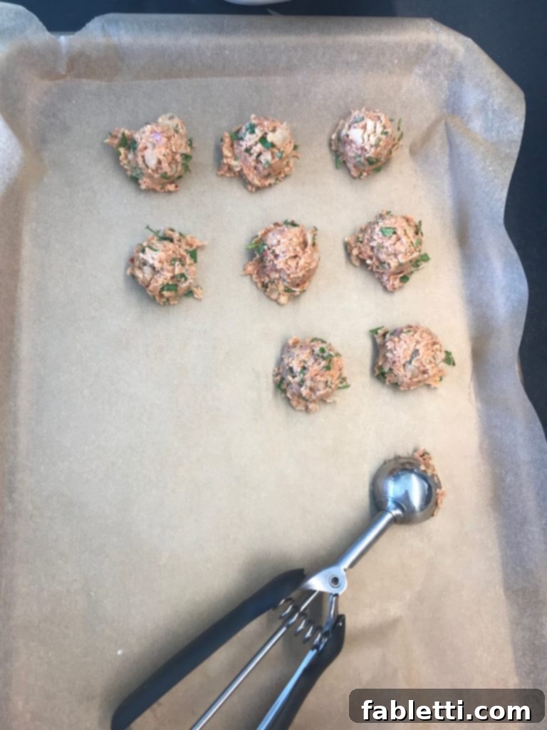 A large rimmed baking tray meticulously lined with unbleached parchment paper, where a small scoop is precisely portioning out rows of uniform vegan meatballs.