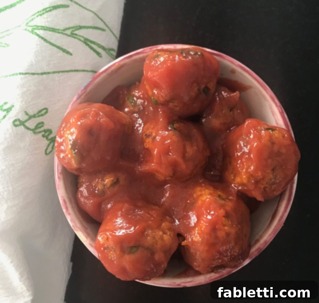 A small, elegant bowl showcasing plump vegan meatballs generously coated in a shimmering sweet and sour sauce, resting beside a white tea towel with a subtle green design. The setup exudes an inviting appetizer vibe.