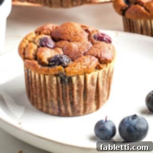 Quick Blender Almond Blueberry Muffins 11 A single blueberry muffin on a white plate, garnished with a few fresh blueberries.