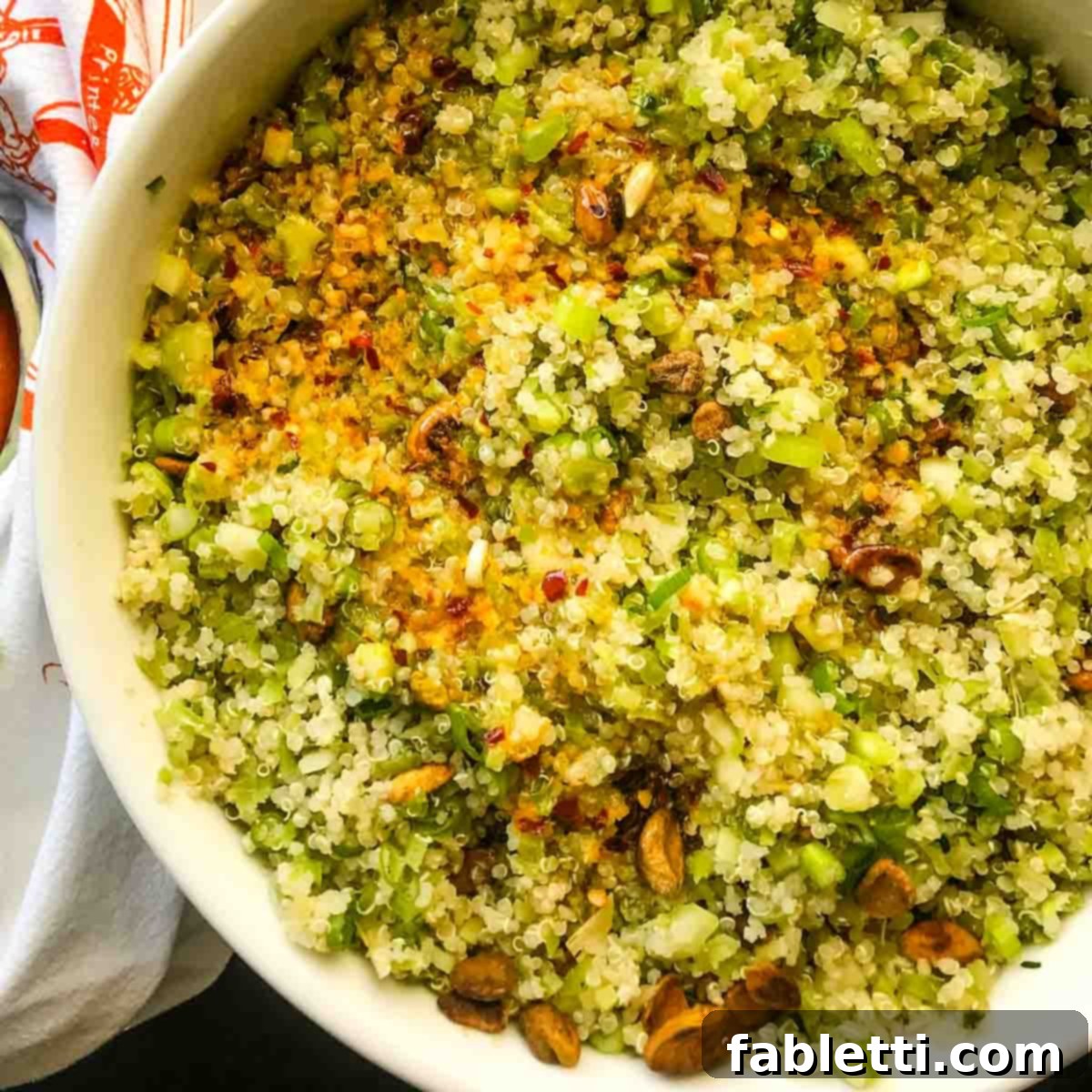 A large bowl of riced broccoli and quinoa salad, with the vibrant orange dressing being poured over, ready to be mixed.