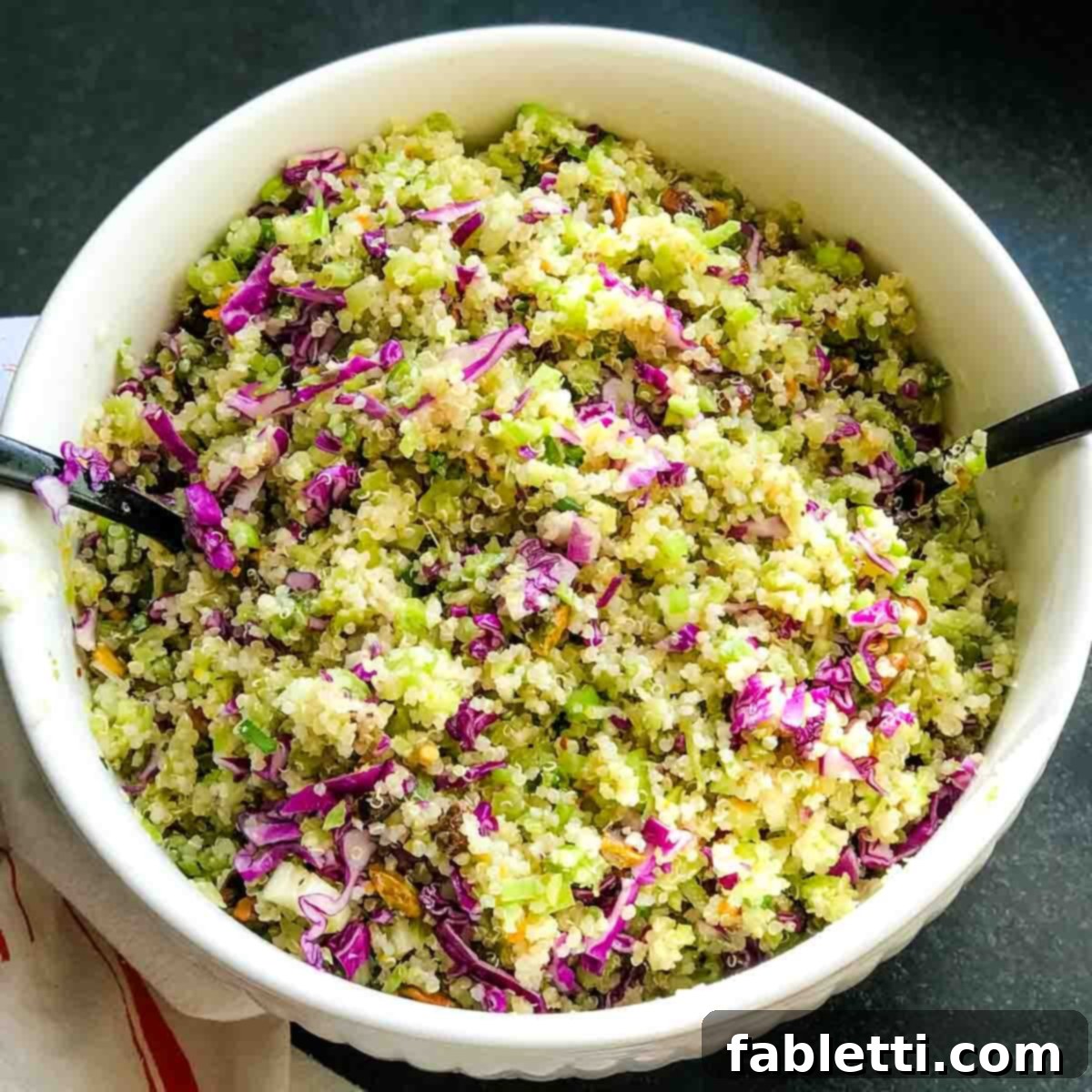 A close-up shot of the raw ingredients for the salad in a bowl: riced broccoli, quinoa, shredded red cabbage, pistachios, and sliced scallions, waiting for the dressing.