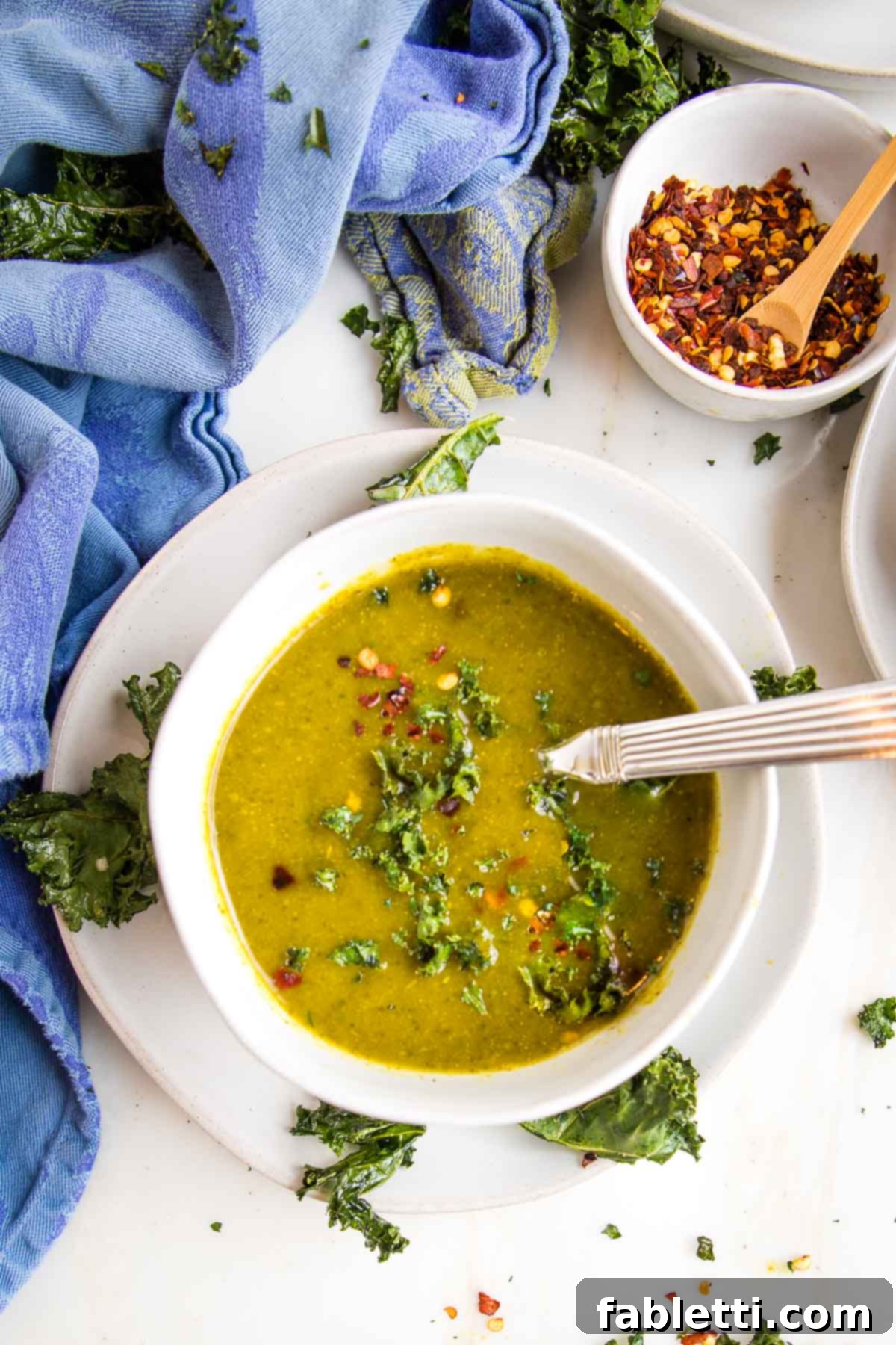 Nourishing Parsnip and Kale Soup 2 Deep green creamy parsnip and kale soup in a white bowl on a white saucer, garnished with crispy kale and red pepper flakes.
