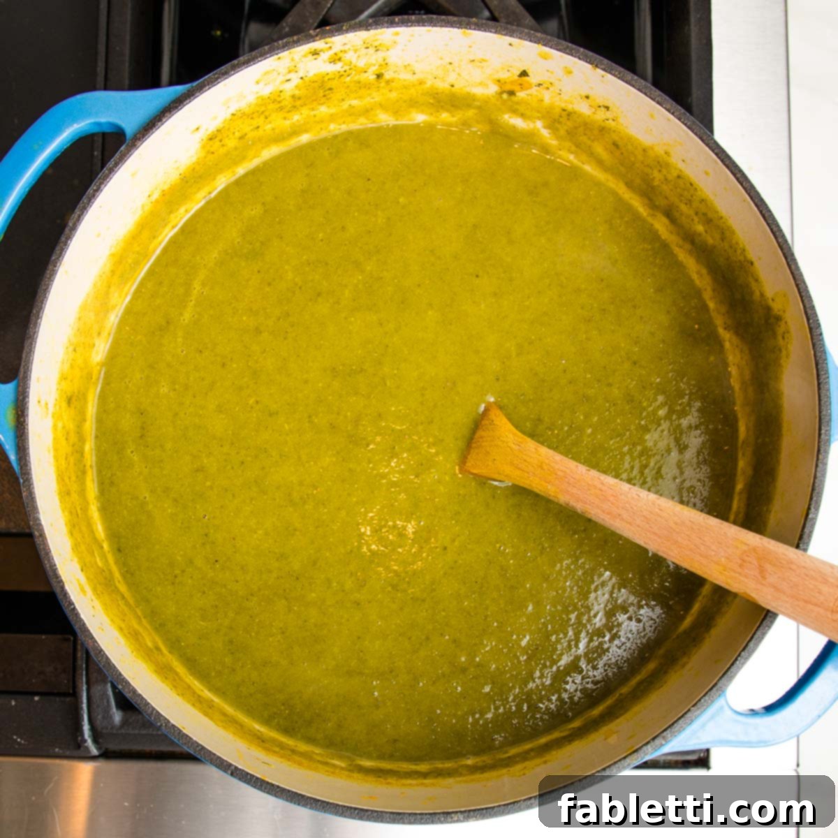 Nourishing Parsnip and Kale Soup 17 Blue Dutch oven on the stove, filled with a pureed green kale soup, with a wooden spoon.