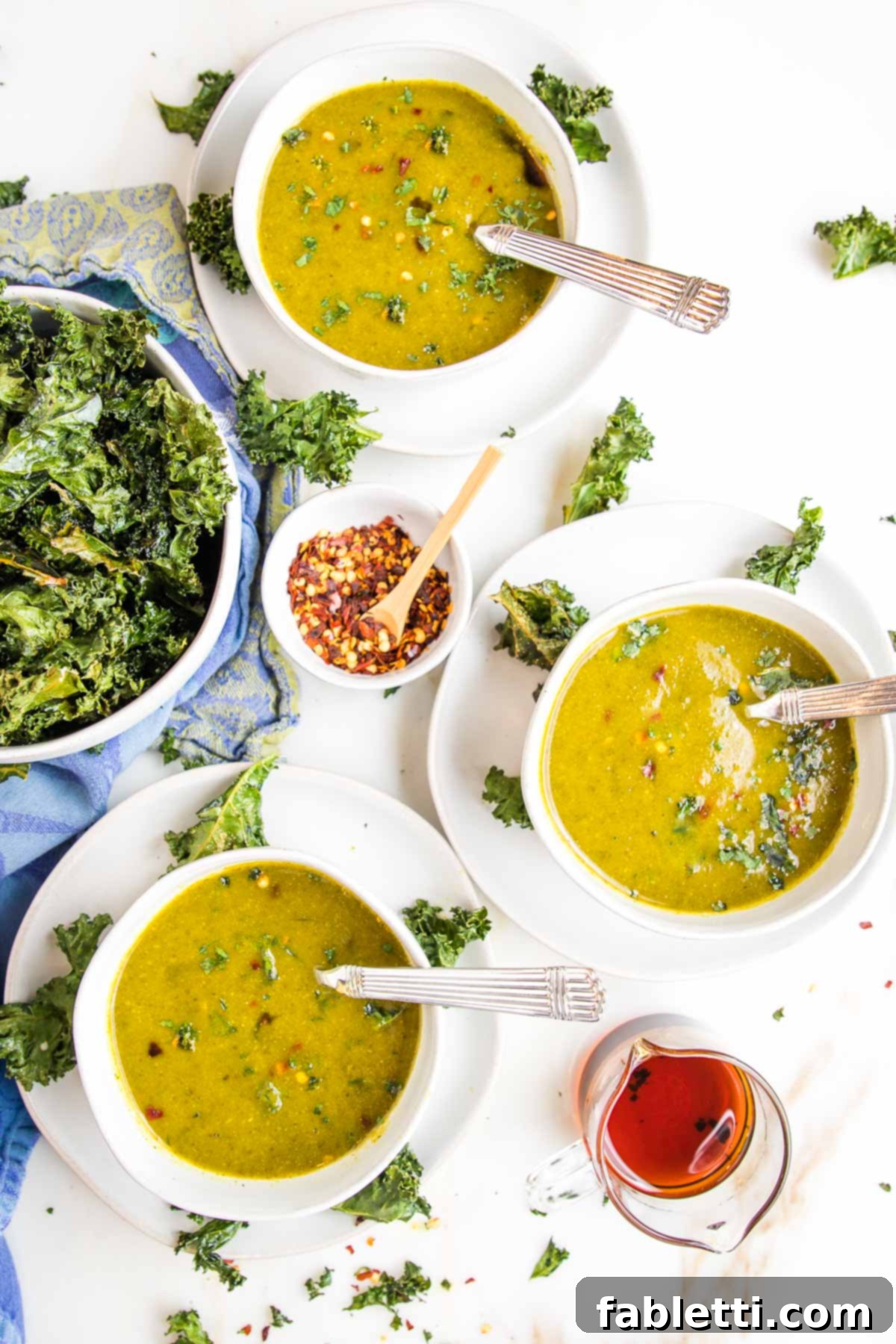 Nourishing Parsnip and Kale Soup 19 Three white bowls filled with creamy kale soup, a bowl of kale chips, and a small dish of crushed red pepper, ready for customization.