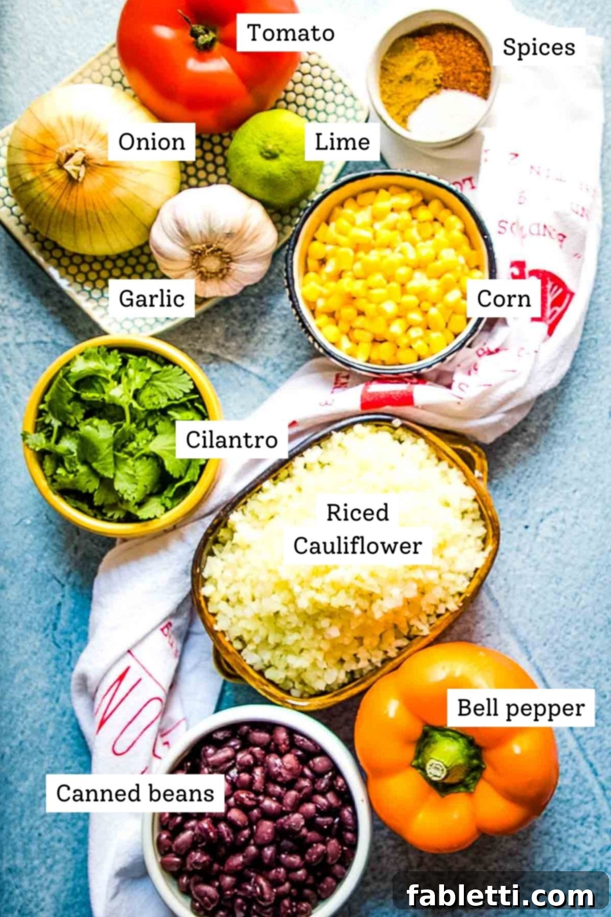 Flavorful Low Carb Mexican Cauliflower Rice 4 A flat lay of labeled ingredients for Mexican cauliflower rice: corn, cilantro, black beans, bell pepper, riced cauliflower, garlic, onions, tomato, spices, and lime.