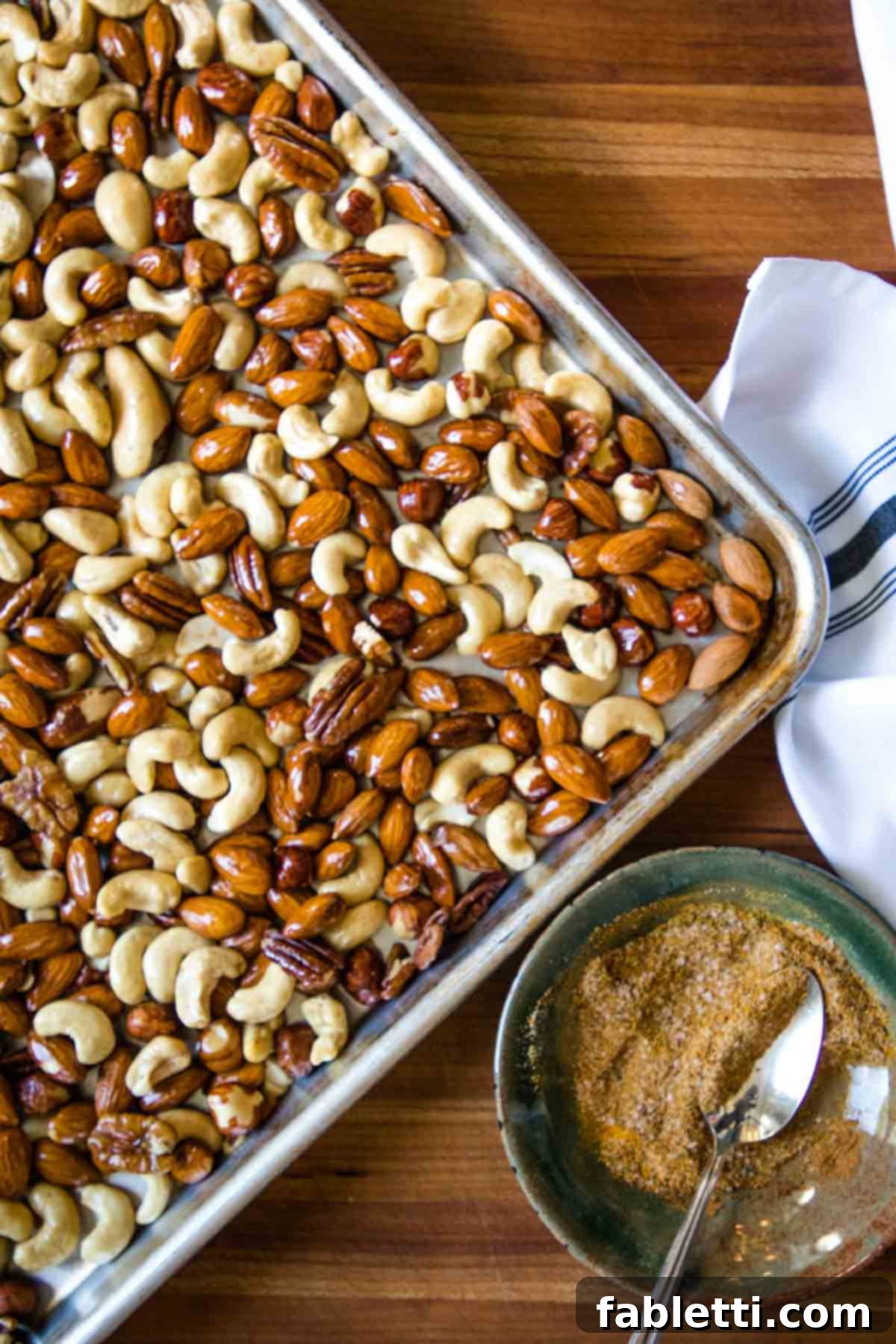 Rimmed baking tray with assorted nuts and a small bowl of spice mixture ready to spoon onto the nuts.
