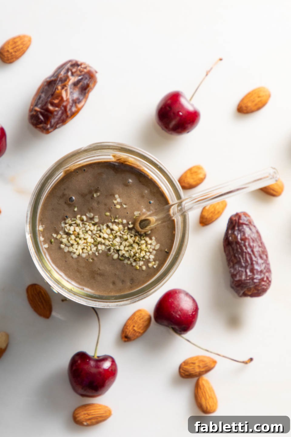 A chocolate smoothie in a glass with hemp seeds sprinkled on top. Fresh dates, cherries, and almonds are artfully arranged on the countertop beside it, hinting at the smoothie's wholesome ingredients.