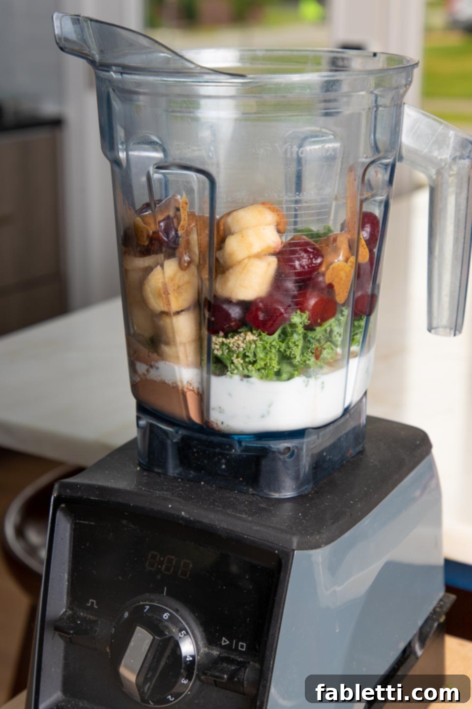 A high-speed blender pitcher is filled with the vibrant ingredients for the chocolate cherry smoothie: chunks of banana, frozen cherries, fresh kale, a splash of almond milk, a dollop of almond butter, rich cacao powder, and a sprinkle of hemp seeds, ready to be blended into a creamy concoction.