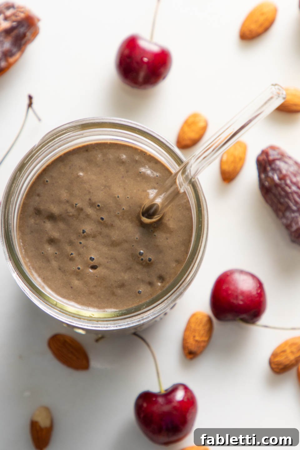A tall glass of rich chocolate cherry smoothie, adorned with a straw, rests on a light countertop. Beside it are scattered fresh cherries, whole almonds, and plump dates, suggesting the wholesome ingredients within.