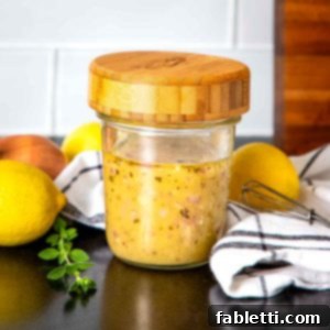Effortless Everyday Salad Dressing 7 Glass jar with wooden lid filled with salad dressing, surrounded by fresh lemons, a shallot and some fresh herbs.