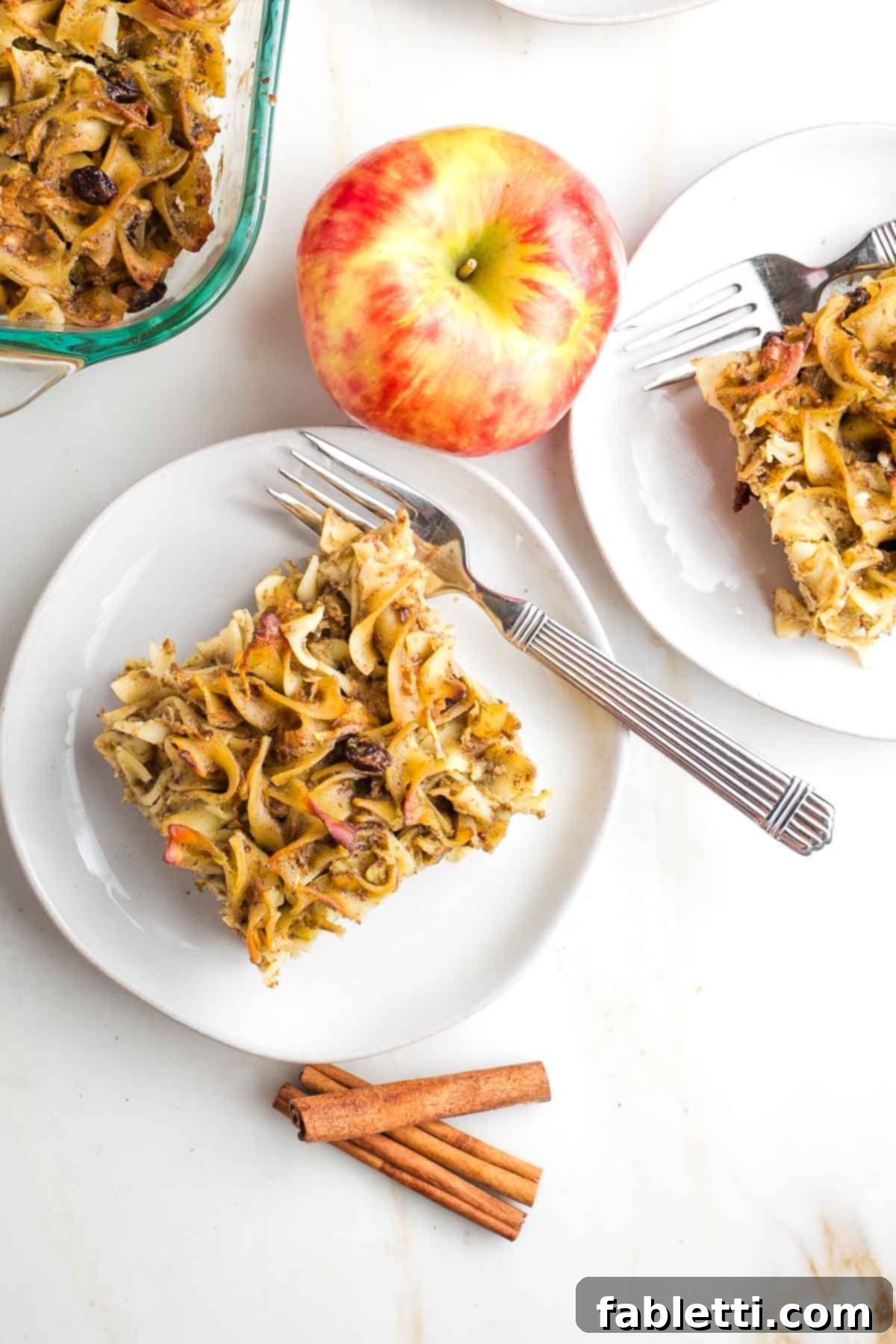 Delicious Dairy-Free Apple Noodle Kugel 2 A delicious piece of apple noodle kugel with raisins sits on a white plate, beautifully garnished with cinnamon sticks and a fresh apple in the soft-focused background, ready to be enjoyed as a sweet side dish or dessert.