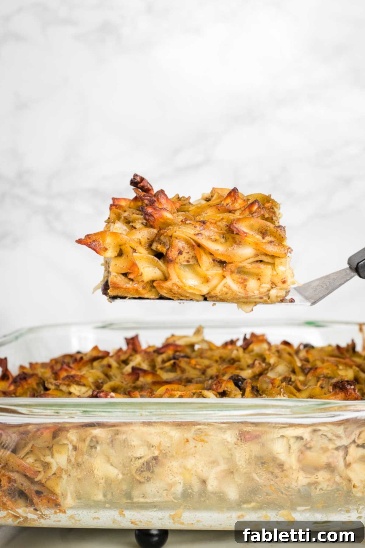Delicious Dairy-Free Apple Noodle Kugel 14 A perfect square of golden-brown apple noodle kugel, dotted with raisins, is gently lifted from the casserole dish with a spatula, showcasing its tender interior and crispy top layer.