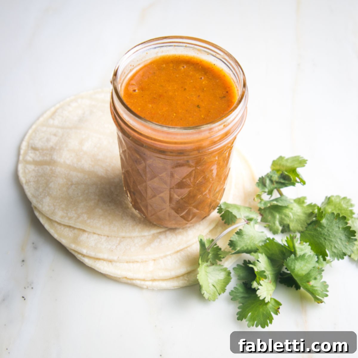Jar of red enchilada sauce with cilantro and tortillas on the side.