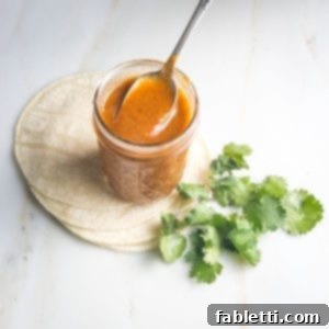 Jar of red enchilada sauce with a spoon in it on top of a stack of tortillas with some cilantro on the side.