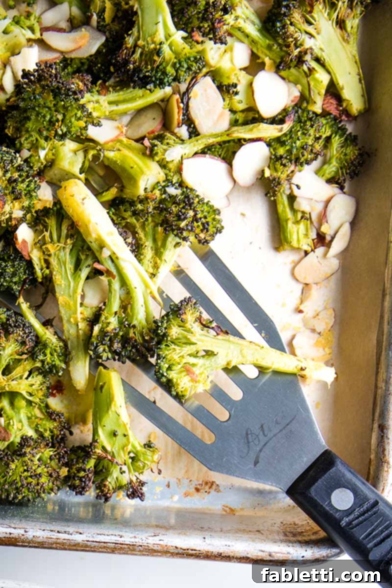 Close-up of golden brown, crispy roasted broccoli with toasted almonds and fresh lemon wedge on a sheet pan.