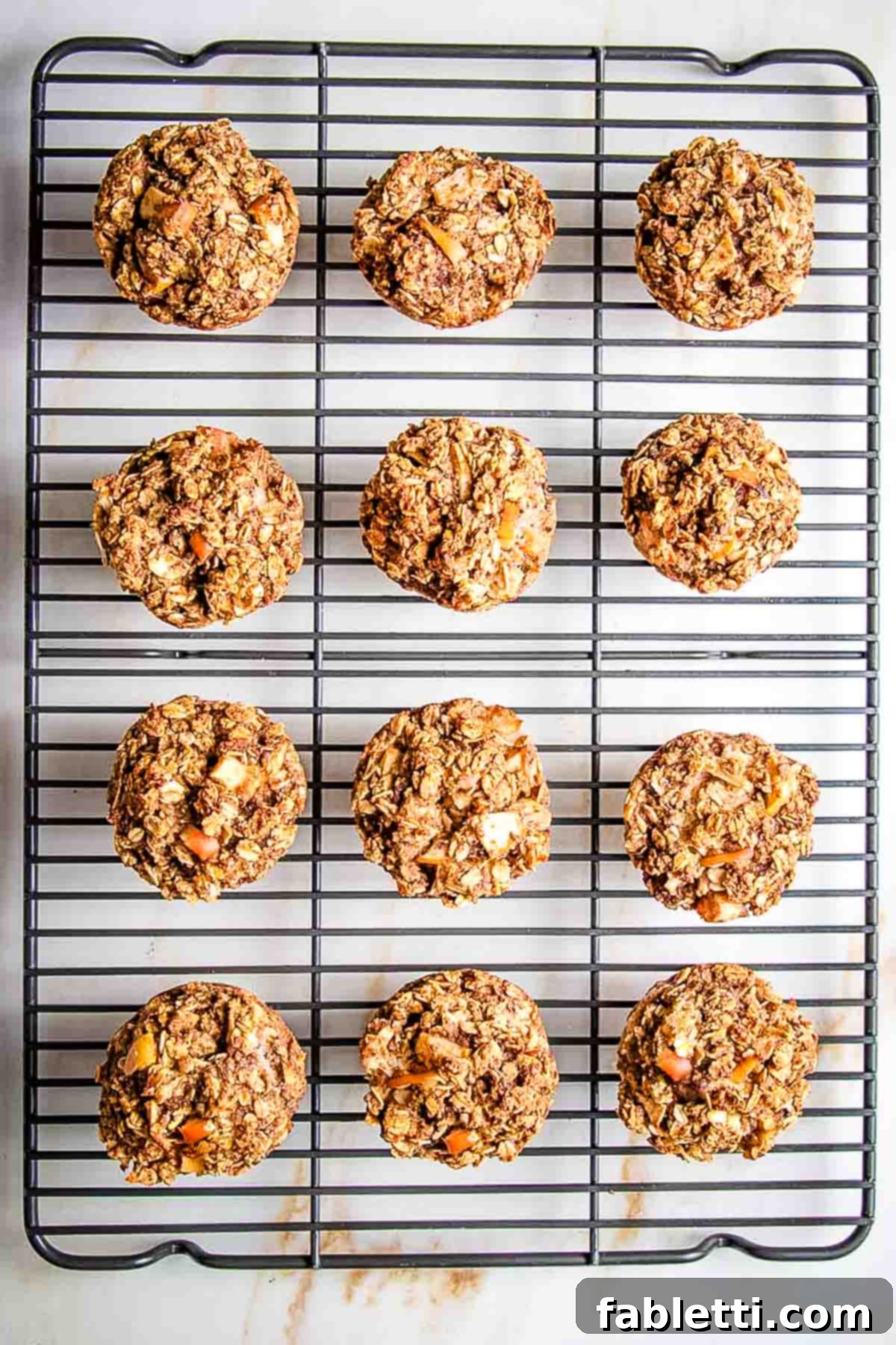 A freshly baked oatmeal apple muffin being lifted out of the tin using parchment strips, then placed on a wire rack.