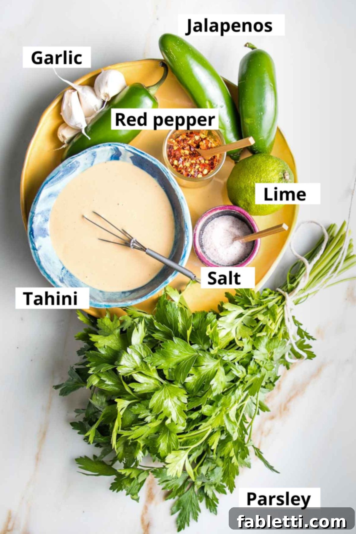Carefully arranged ingredients for green tahini sauce on a wooden board: rich tahini, vibrant jalapenos, fresh lime, pungent garlic, fragrant parsley, essential salt, and a pinch of red pepper flakes.