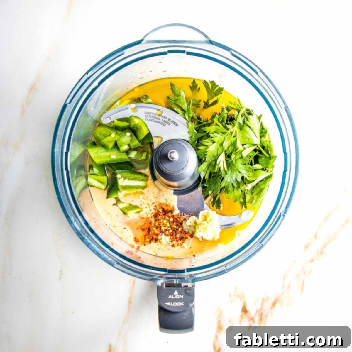 A food processor bowl filled with the fresh ingredients for green tahini sauce: smooth tahini, vibrant jalapeno slices, fresh parsley leaves, a sprinkle of crushed red pepper, and finely grated garlic, ready to be blended.