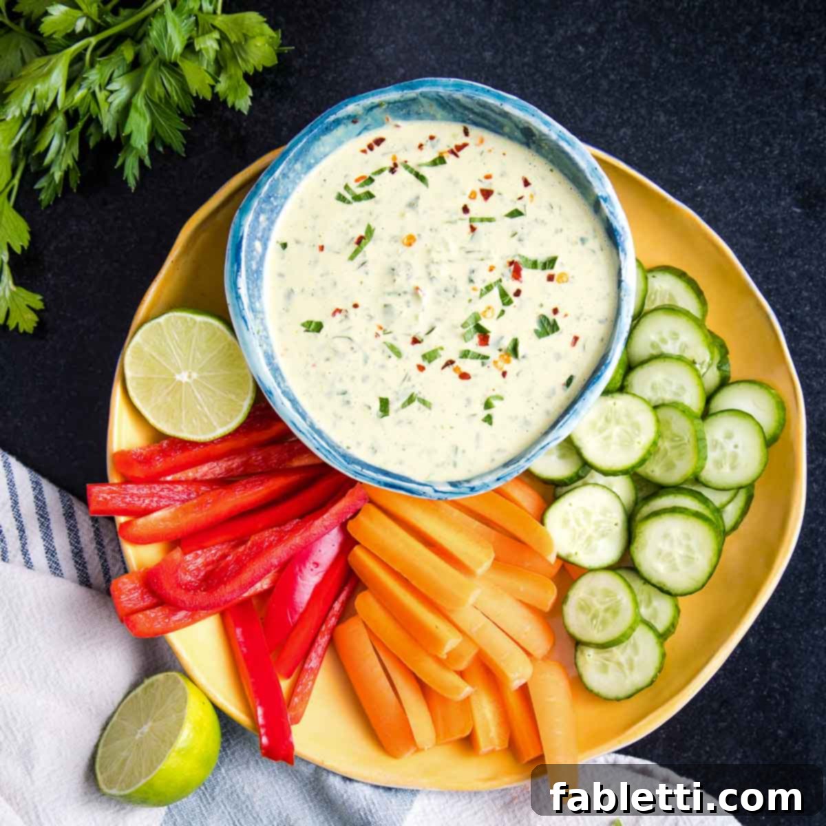 A inviting bowl of green tahini sauce is centrally placed, surrounded by a vibrant selection of neatly sliced peppers, crisp carrots, and refreshing cucumbers, artfully arranged on a clean white plate, creating a perfect setup for dipping.