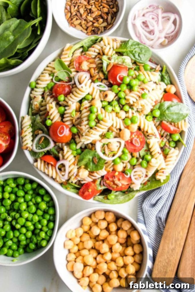 No-Fuss Summer Pasta Salad 2 Small white bowls with chickpeas, green peas, halved cherry tomatoes, spinach, sunflower seeds, and sliced shallots arranged around a larger white bowl of pasta salad, showcasing the fresh ingredients.