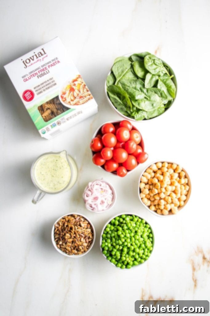 No-Fuss Summer Pasta Salad 3 Top-down view of essential ingredients: a box of gluten-free fusilli pasta, a pitcher of green tahini sauce, and small white bowls filled with fresh spinach, cherry tomatoes, sliced shallots, shelled sunflower seeds, green peas, and chickpeas.