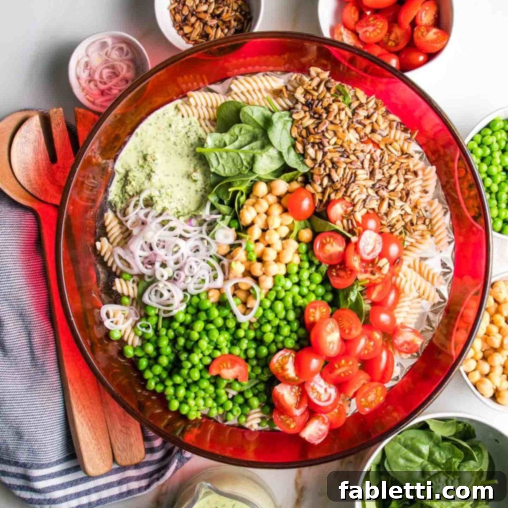 No-Fuss Summer Pasta Salad 4 A large bowl with a red rim is filled with pasta, generously topped with vibrant red cherry tomatoes, piles of green peas, a handful of chickpeas, a drizzle of green tahini sauce, and a sprinkle of sunflower seeds, ready to be mixed.