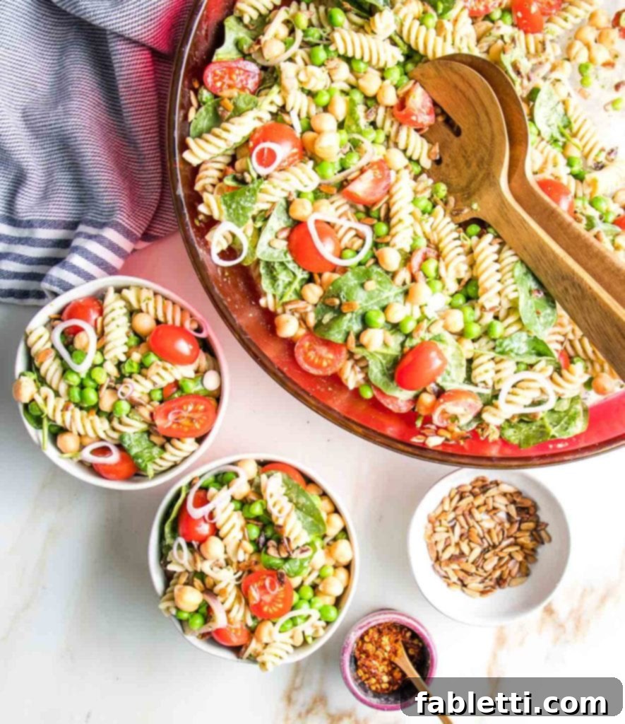 No-Fuss Summer Pasta Salad 5 A large red glass bowl of colorful pasta salad is being served into smaller white bowls, with extra sunflower seeds and red pepper flakes available for garnish.