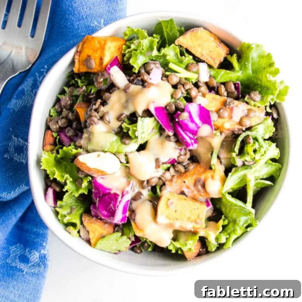 Vibrant Vegan Lentil Medley 15 A white bowl filled with kale, cabbage, sweet potato cubes, black lentils, onions, almonds, parsley, and a creamy dressing, ready to be enjoyed.