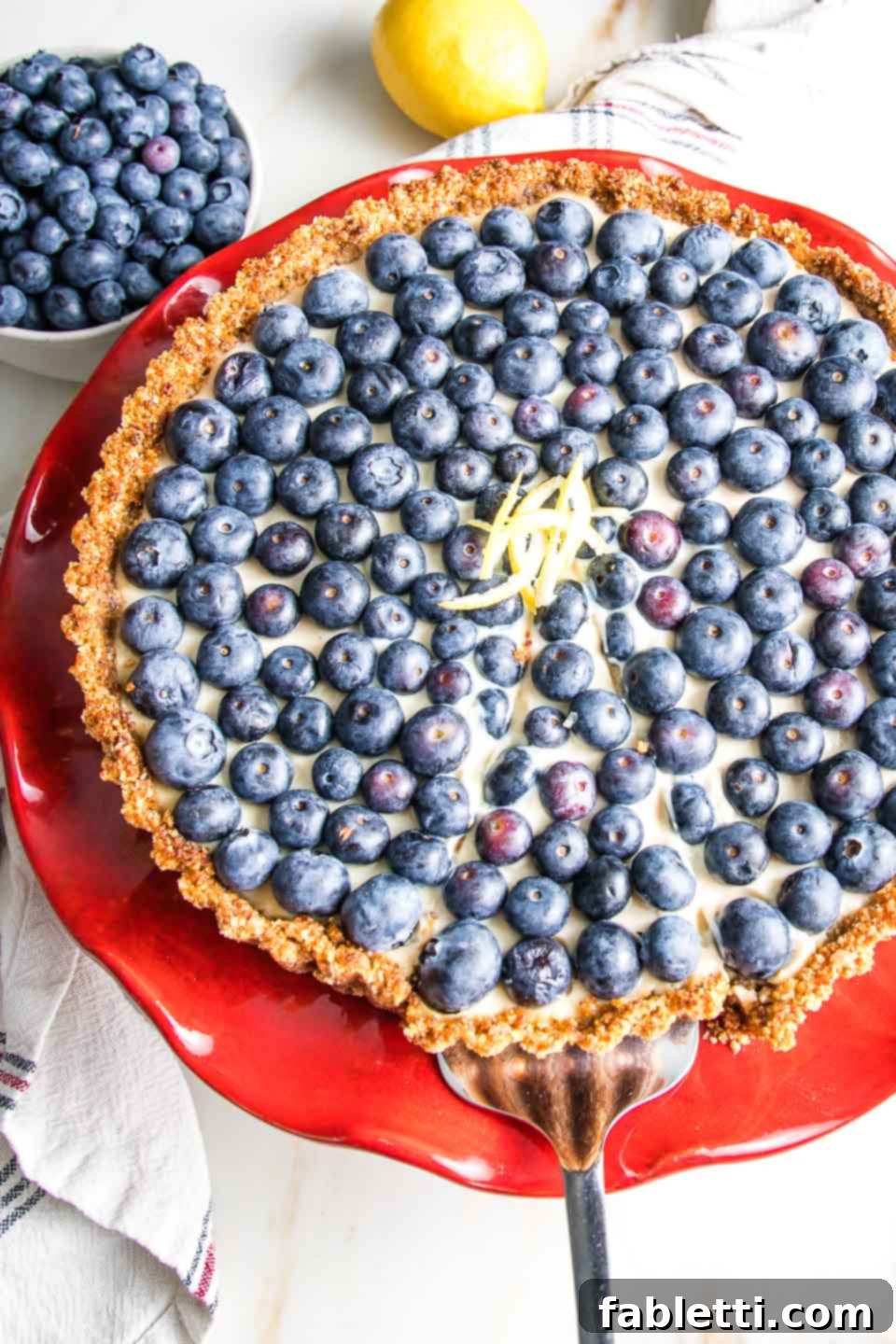 Homemade Blueberry Tart Perfection 2 Vibrant blueberry tart on a red cake stand, elegantly garnished with lemon peel. A backdrop of fresh lemons and a bowl of blueberries completes the inviting scene.