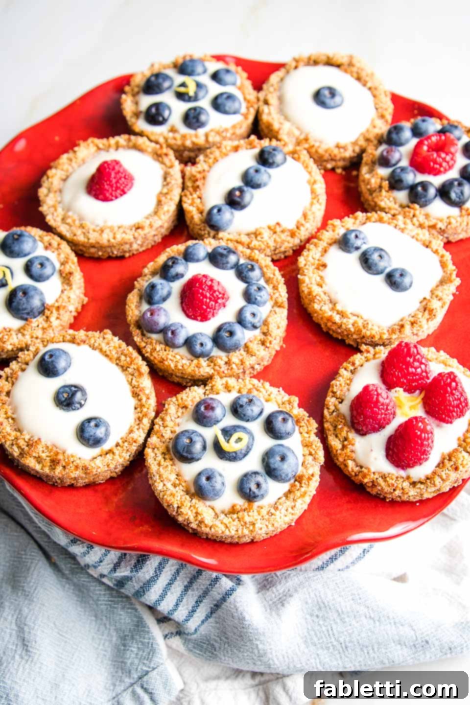 Homemade Blueberry Tart Perfection 11 A bright red cake stand holding an array of mini berry tarts, artfully decorated with both raspberries and blueberries, ready for serving.
