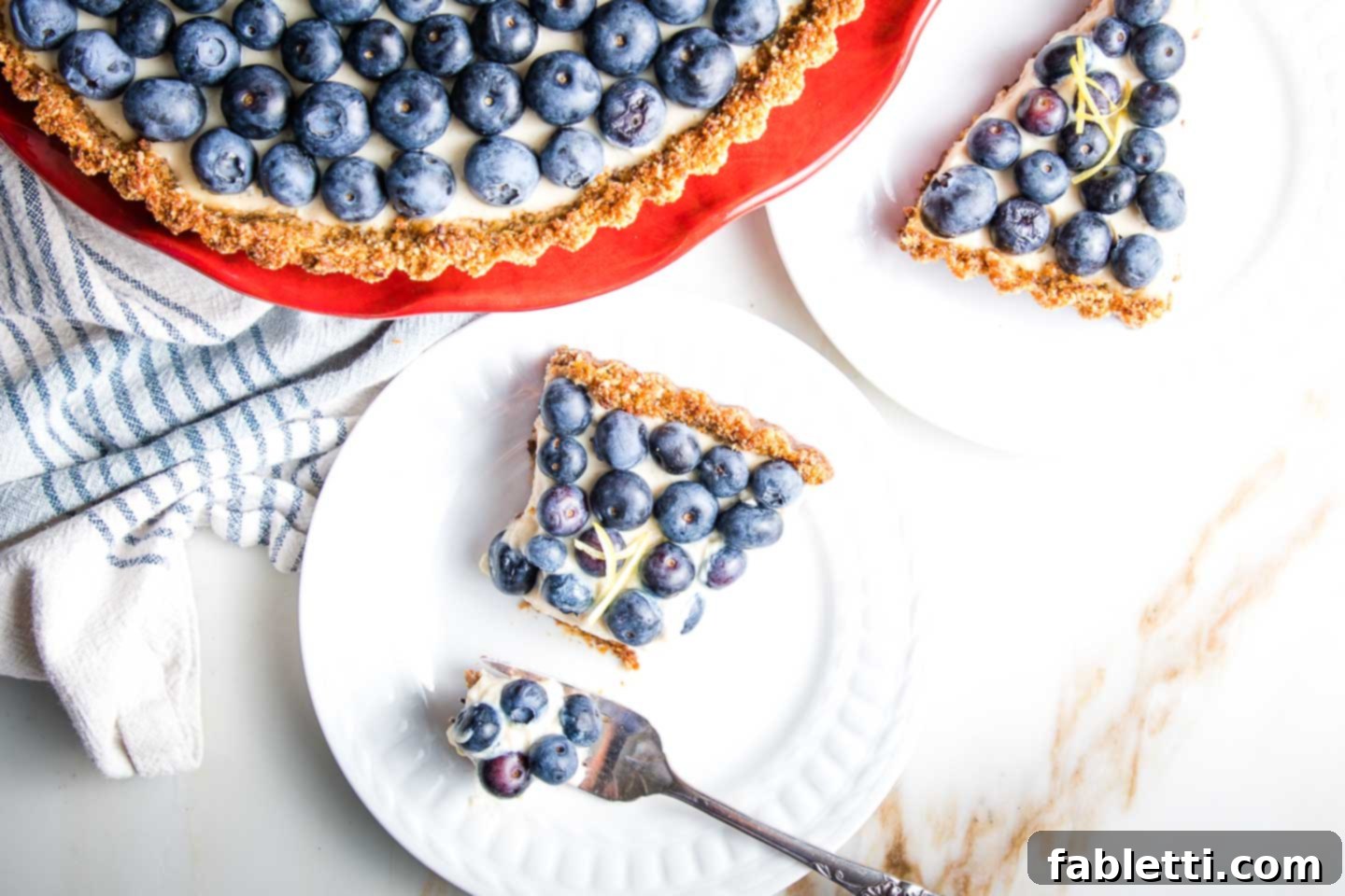 Homemade Blueberry Tart Perfection 3 A perfectly portioned slice of blueberry tart rests on a white plate, positioned next to a red cake stand holding the full tart. The tart showcases a beautiful browned crust, a creamy white filling, and a generous topping of fresh blueberries.
