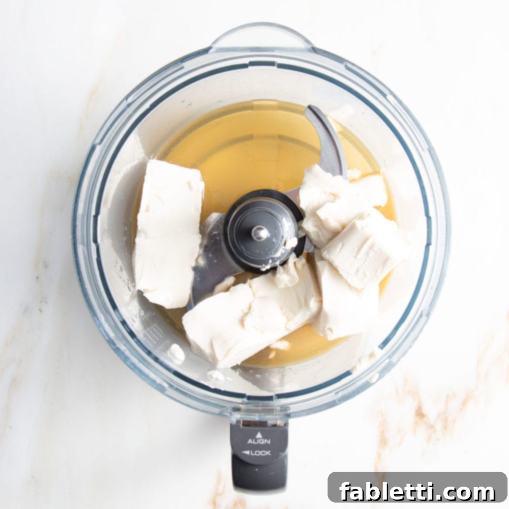 Filling for healthy blueberry tart is easy to make in the food processor. Tofu, maple syrup, lemon juice and vanilla extract, ready to process for debra klein recipe. A food processor bowl containing silken tofu, maple syrup, lemon juice, and vanilla extract, poised to be blended into a creamy vegan filling for a healthy blueberry tart.