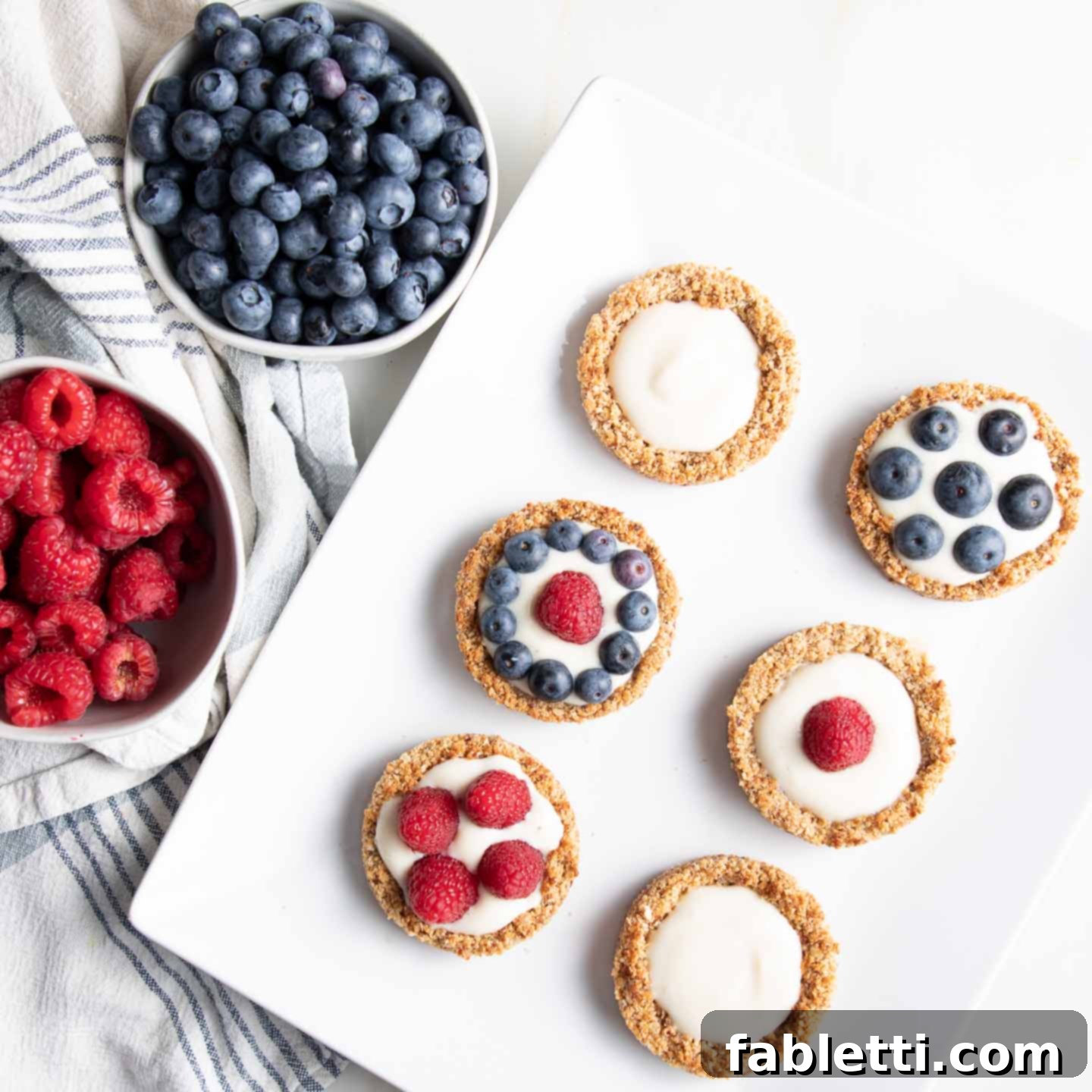 Homemade Blueberry Tart Perfection 9 A vibrant display featuring bowls of fresh blueberries and raspberries, accompanied by a platter of mini blueberry tarts. Some tarts are filled with the creamy white tofu filling, while others are generously topped with a mix of fresh berries.
