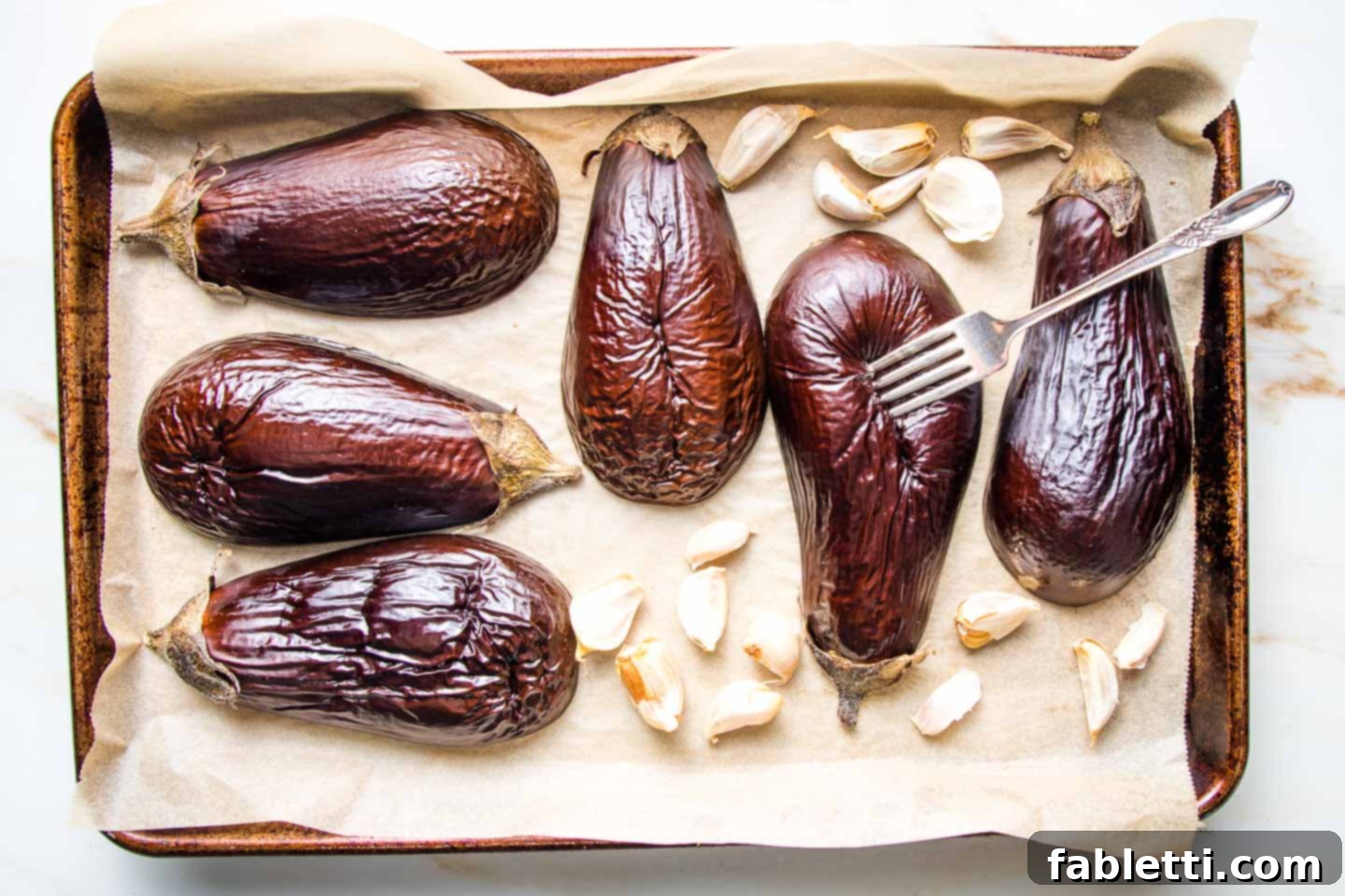 Velvety Roasted Garlic and Tahini Baba Ganoush 5 Halved eggplant roasting on a parchment lined baking tray with garlic cloves in their skins.