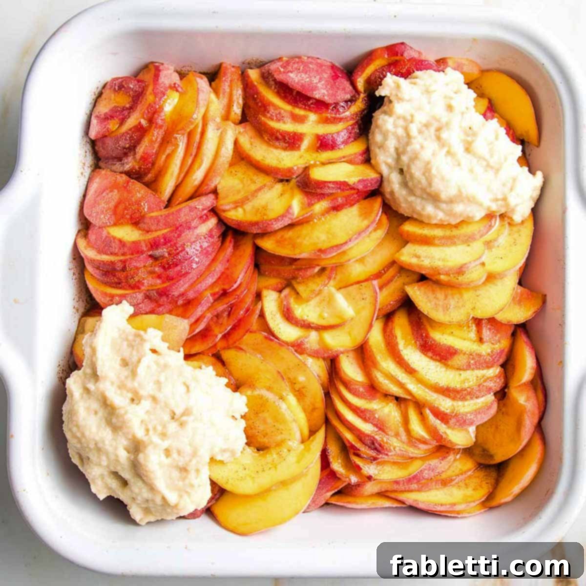 Sliced peaches with a couple dollops of cobbler topping in a square baking dish, ready for more topping.